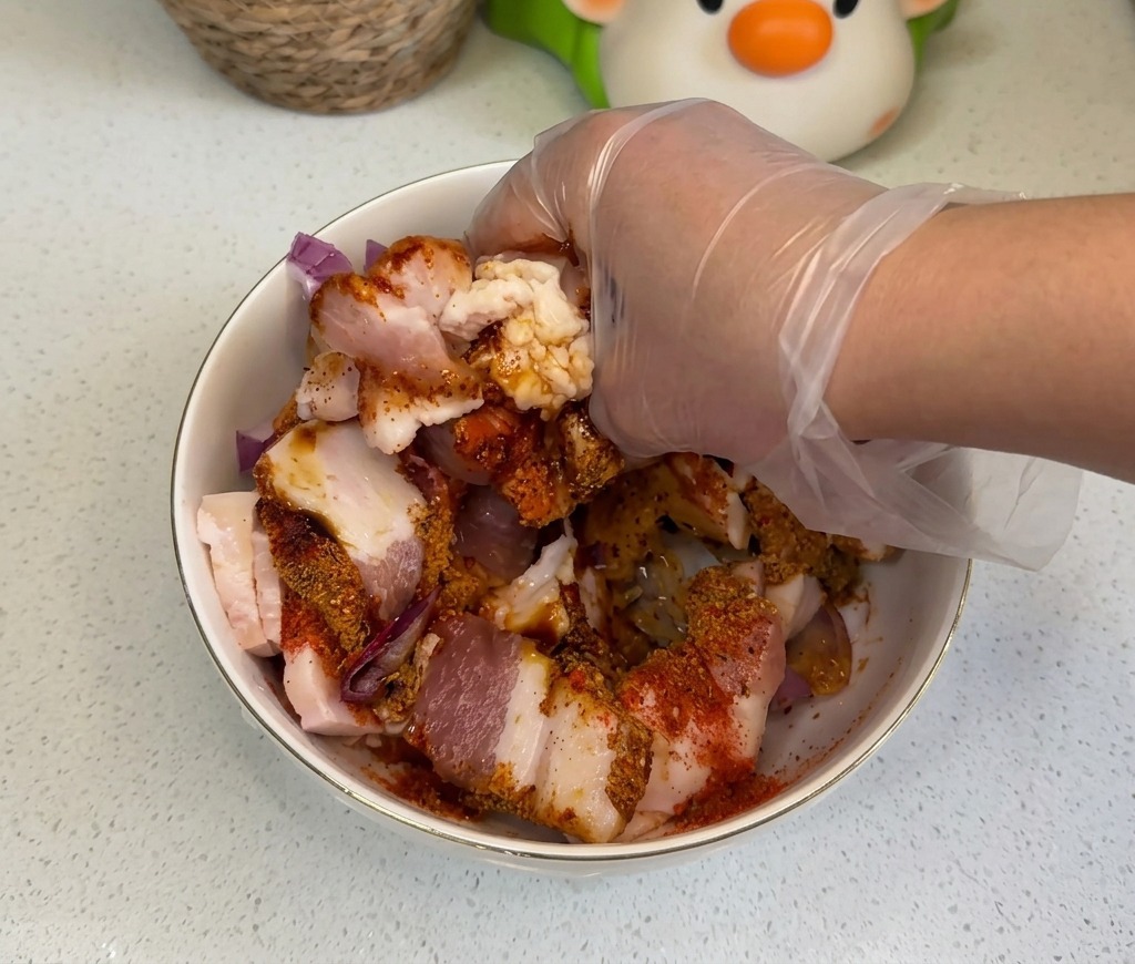 Eine Hand in einem transparenten Plastikhandschuh vermengt rohe Schweinebauchscheiben gründlich mit Zwiebeln und bunten Gewürzen in einer Schüssel.