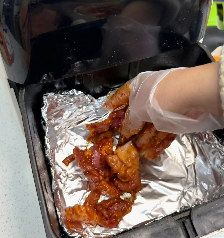 Eine Hand mit einem transparenten Plastikhandschuh legt rot marinierte Schweinebauchscheiben auf silberne Alufolie in einem schwarzen Heißluftfritteusenkorb.