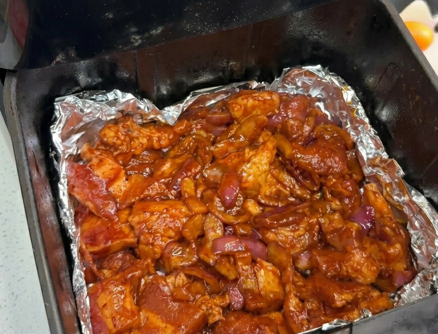 Marinierter Schweinebauch und rote Zwiebeln sind gleichmäßig auf dem mit Folie ausgelegten Boden eines Heißluftfritteusenkorbs verteilt.