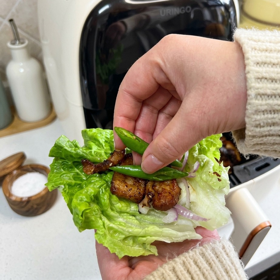 Eine Hand legt zwei kleine, leuchtend grüne Chilischoten auf einen Salatwrap mit knusprigem Schweinebauch und Zwiebeln.