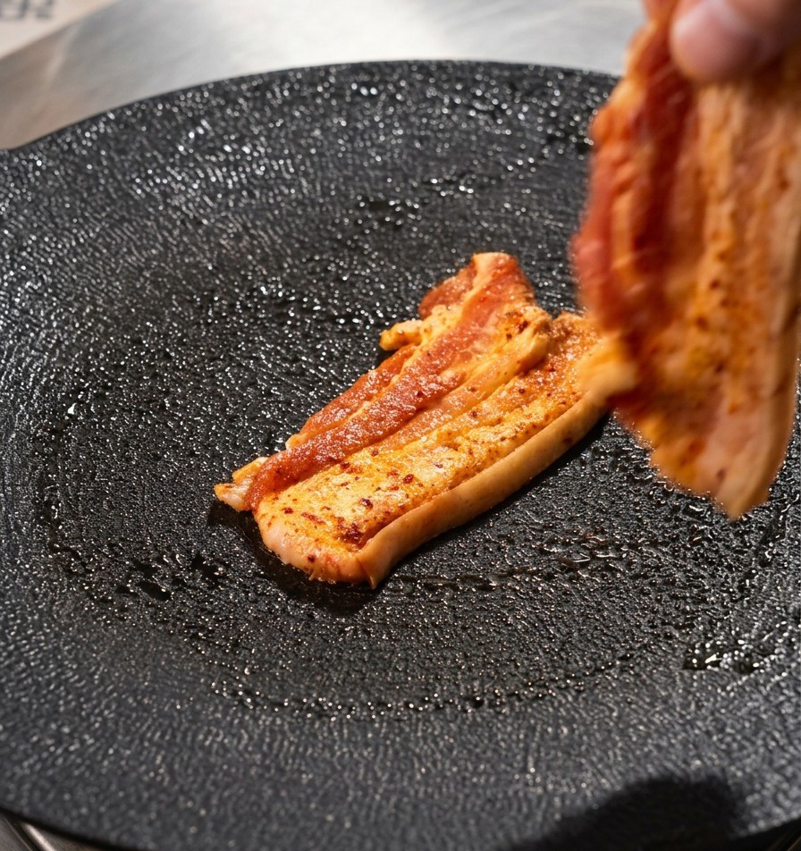 Eine Hand legt einen Streifen Schweinebauch auf eine strukturierte, schwarze, runde Grillpfanne.