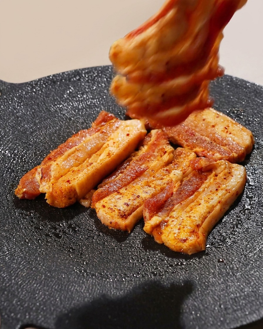 Mehrere Streifen gewürzter Schweinebauch werden nebeneinander auf einer heißen Grillpfanne platziert.