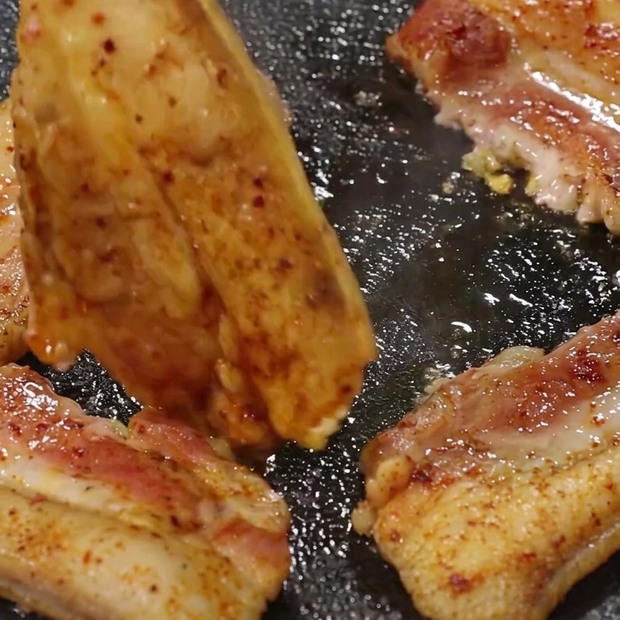 Eine Grillzange wendet ein Stück gebräunten Schweinebauch auf einer heißen, von Fett zischenden Grillpfanne.