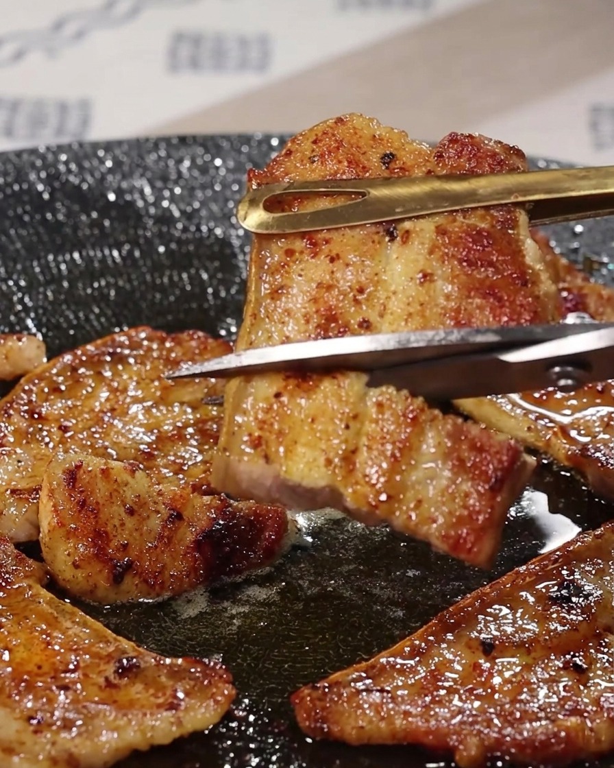 Eine Küchenschere aus Metall schneidet ein Stück gegrillten Schweinebauch, das mit einer Zange über einer zischenden Pfanne gehalten wird.