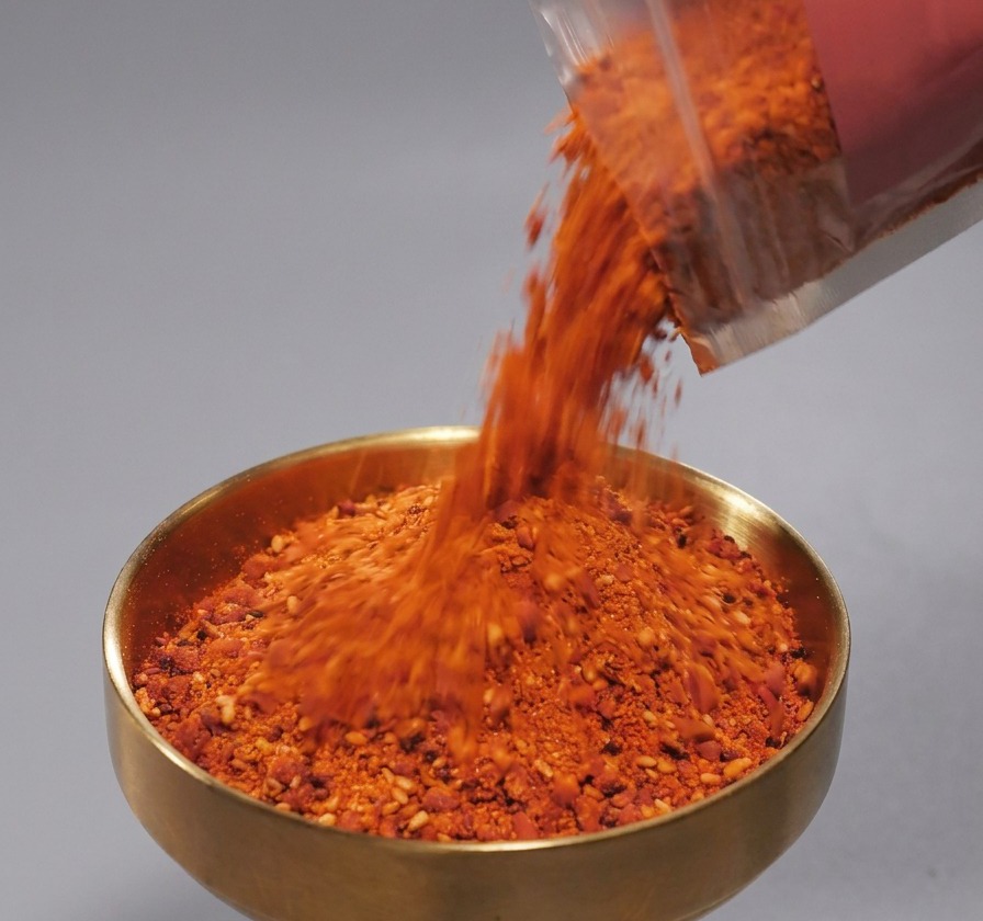 Rotes Gewürzpulver mit Sesamkörnern wird aus einer Kunststoffpackung in ein messingfarbenes Schälchen geschüttet.