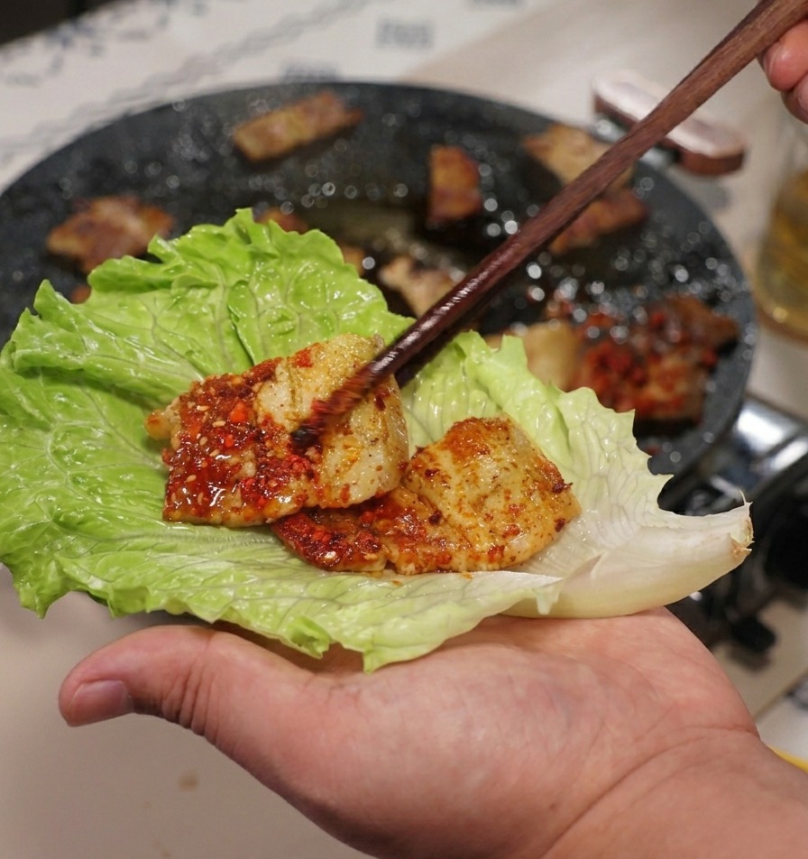Eine Hand hält ein großes, grünes Salatblatt, während mit Stäbchen ein Stück gegrillter Schweinebauch darauf platziert wird.