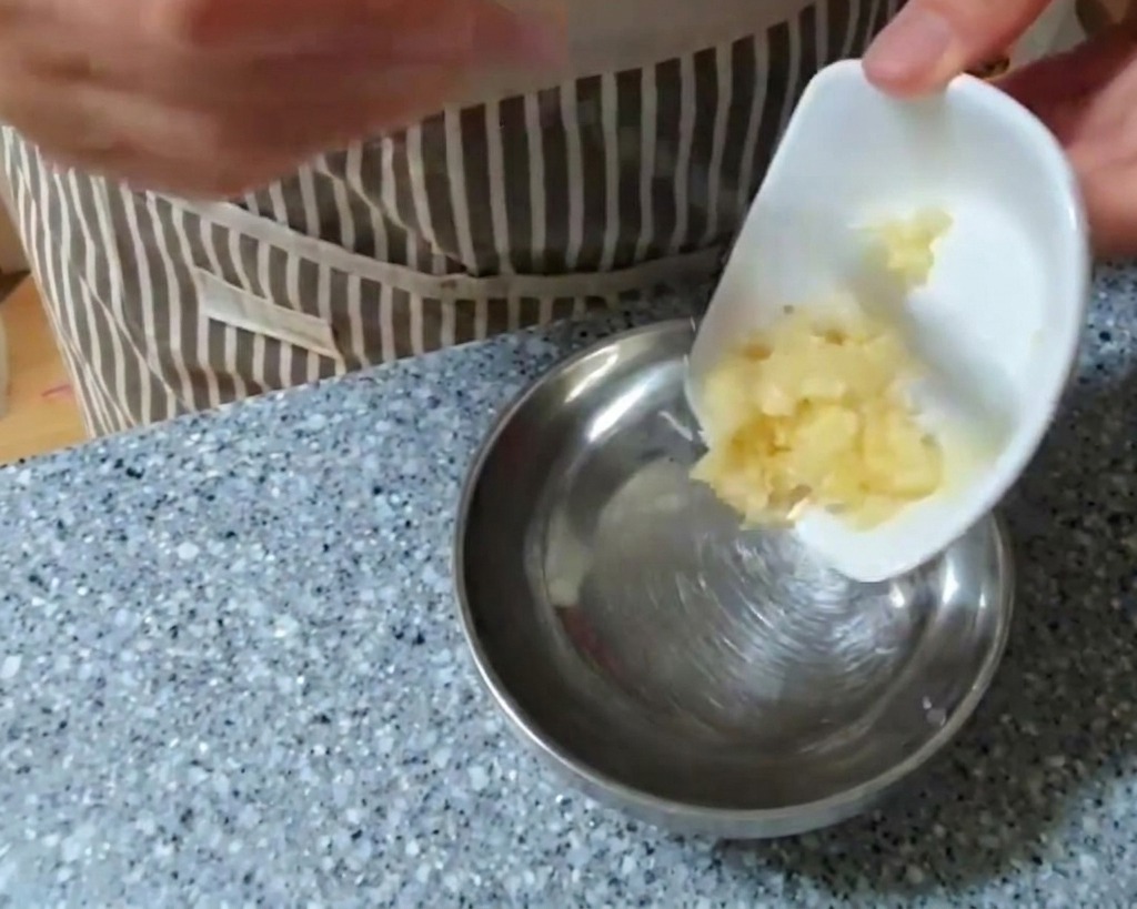 Eine Person gibt eine Portion gehackten Knoblauch aus einer kleinen weißen Schale in eine größere Rührschüssel aus Edelstahl.