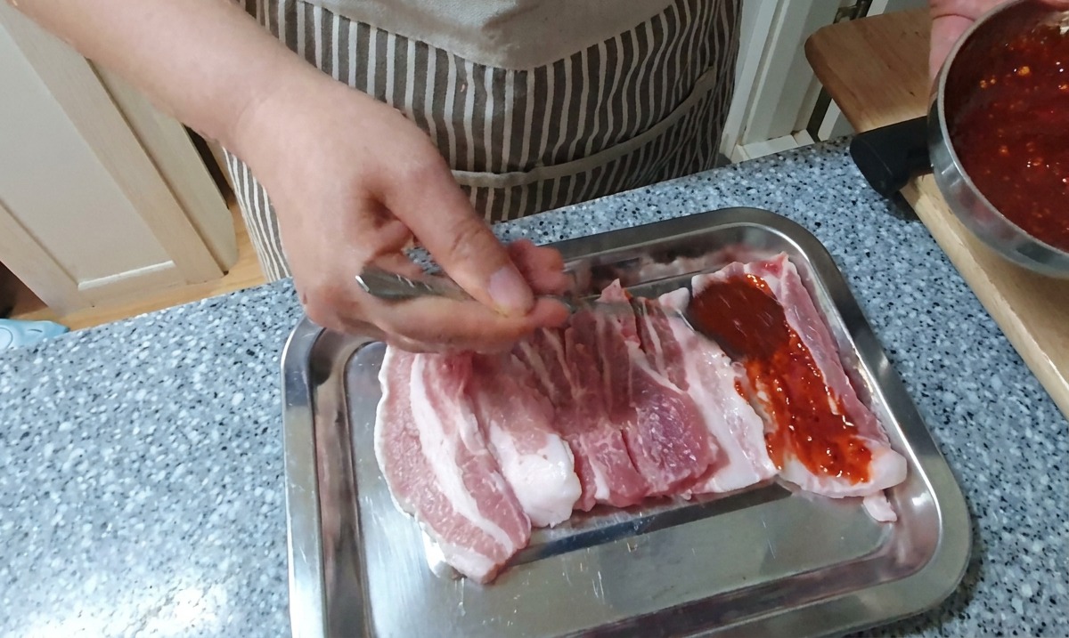 Eine Hand verteilt mit einem Löffel eine dicke, leuchtend rote Chilimarinade auf rohen Schweinebauchscheiben, die auf einem Silbertablett liegen.