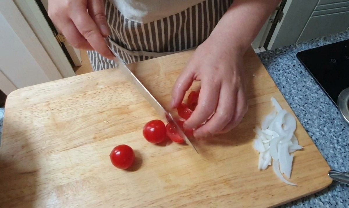 Kleine rote Kirschtomaten werden auf einem Holzbrett neben einem Haufen geschnittener weißer Zwiebeln halbiert.