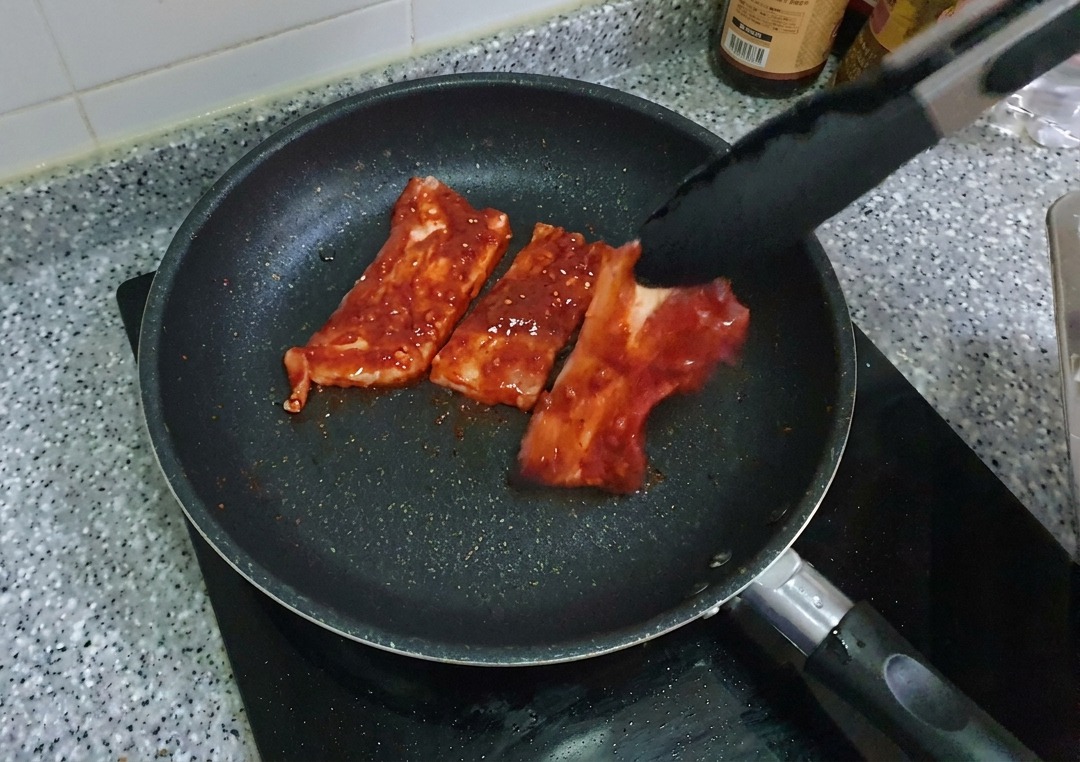 Marinierte, rote Schweinebauchstreifen werden mit einer Grillzange in eine vorgeheizte, schwarze, beschichtete Bratpfanne gelegt.