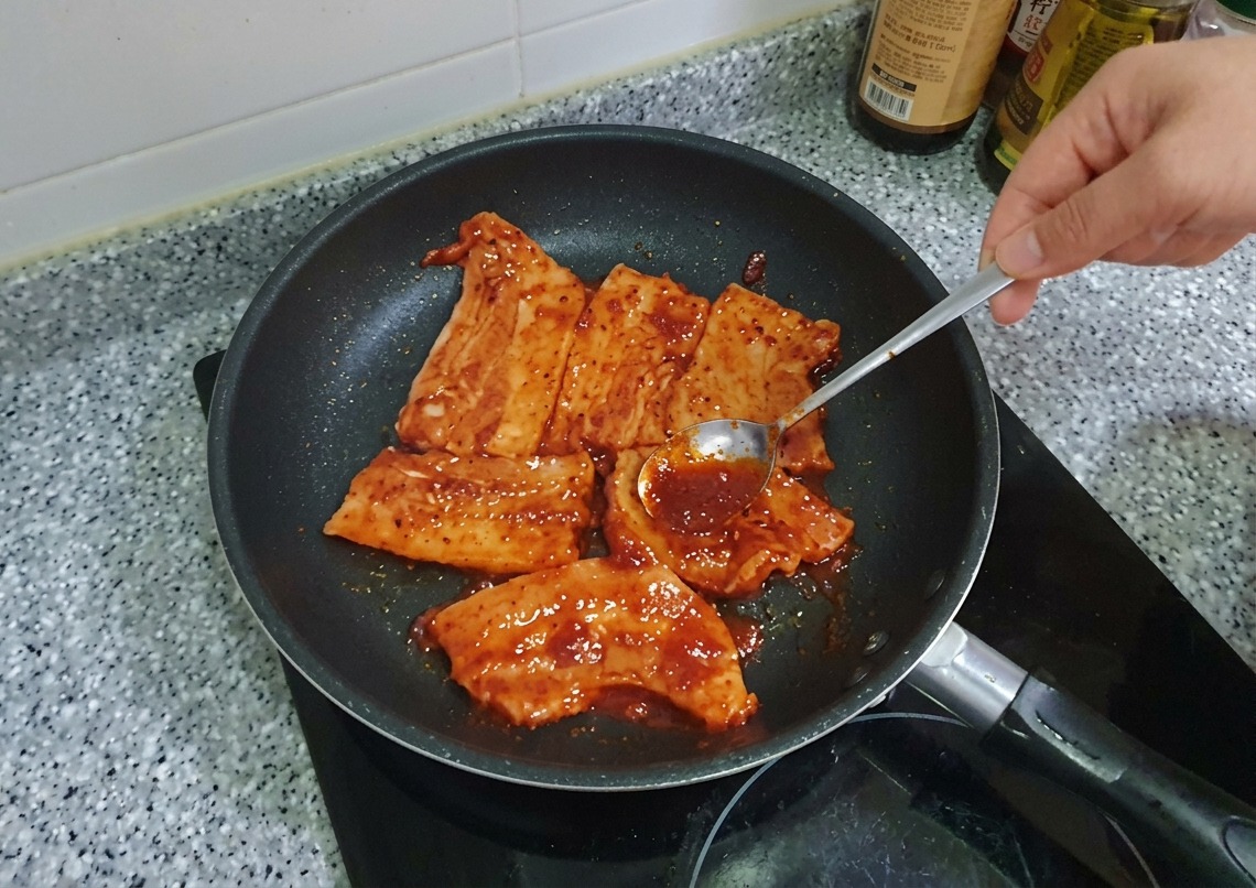 Ein Löffel verteilt dicke, rote Gochujang-Marinade auf den Schweinebauchscheiben in der Pfanne.