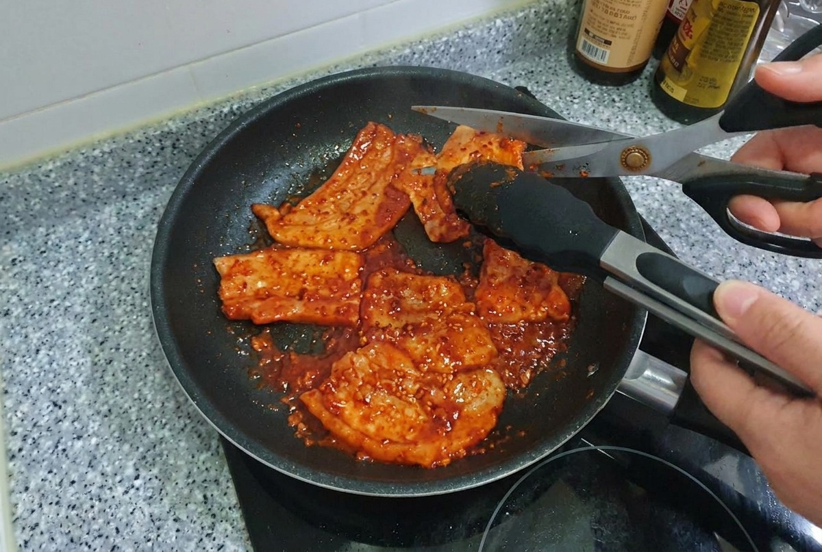 Eine Küchenschere schneidet Streifen von scharfem, gegrilltem Schweinebauch direkt in der Bratpfanne in Stücke.