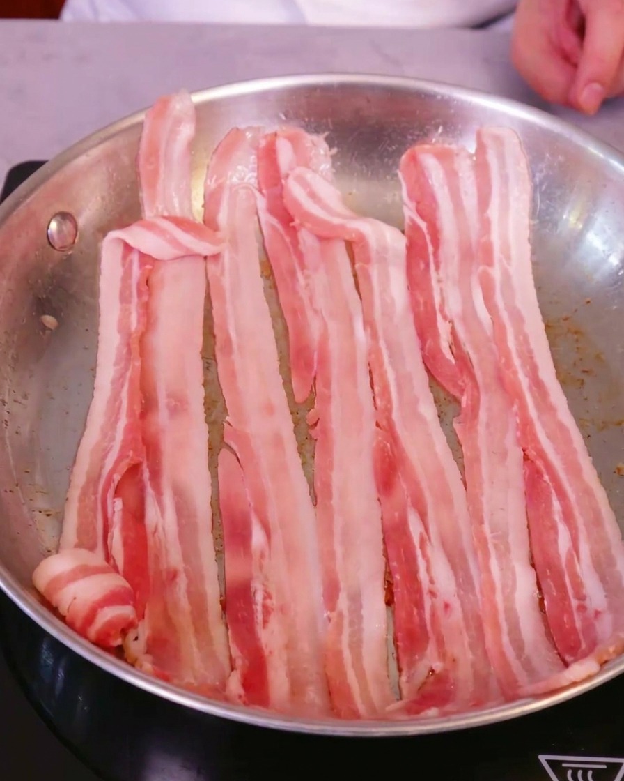 Frische, rohe Schweinebauchscheiben, ordentlich nebeneinander in einer großen Edelstahlpfanne auf einem Induktionsherd angeordnet.
