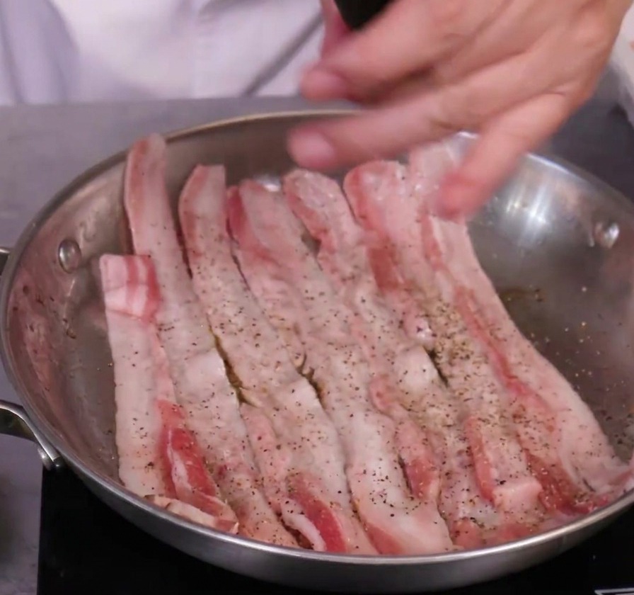 Rohe Schweinebauchscheiben in einer Edelstahlpfanne, die mit einer manuellen schwarzen Pfeffermühle gewürzt werden.