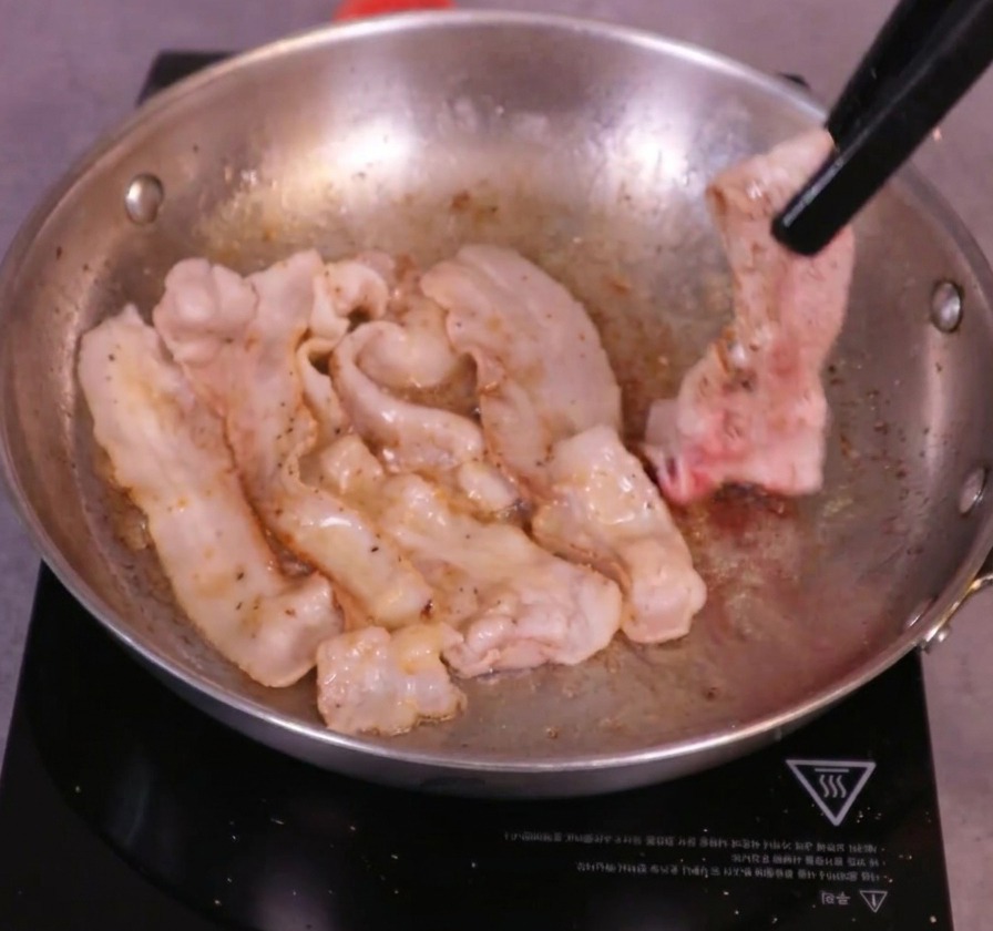 Goldbraune Schweinebauchscheiben, die mit einer schwarzen Küchenzange in einer brutzelnden Pfanne gewendet werden.