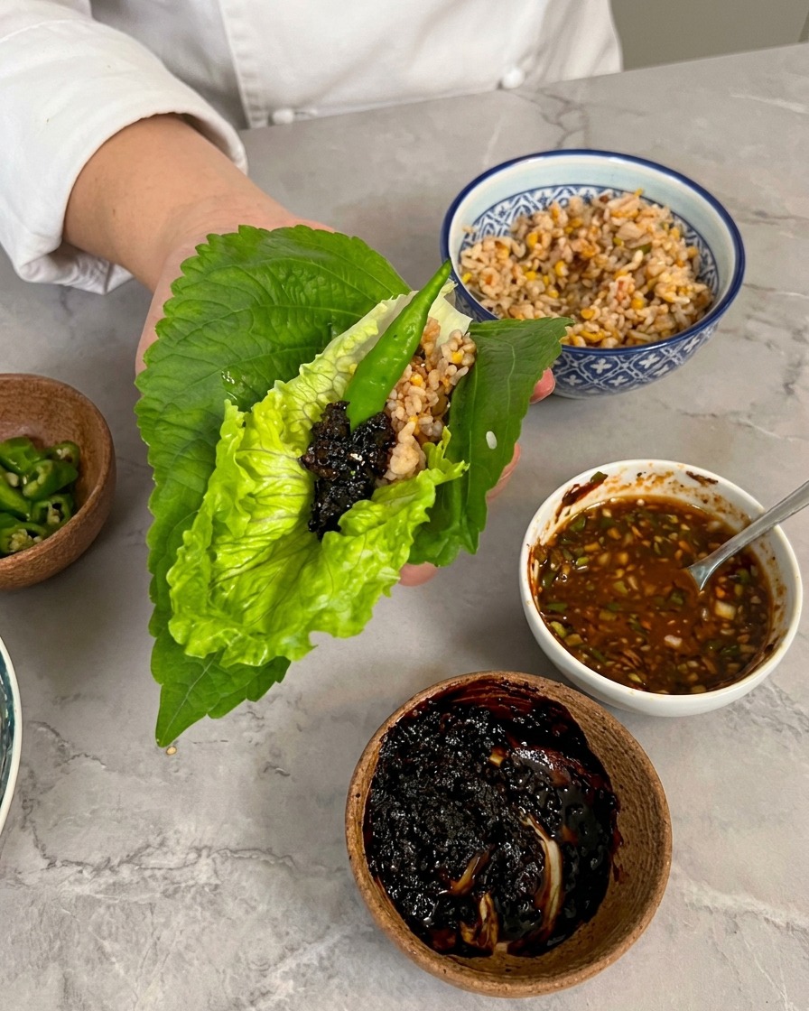 Eine Hand hält einen Wrap aus Salat und Perillablatt, garniert mit einem Löffel dunkler Barbecuesauce und einer grünen Chili.