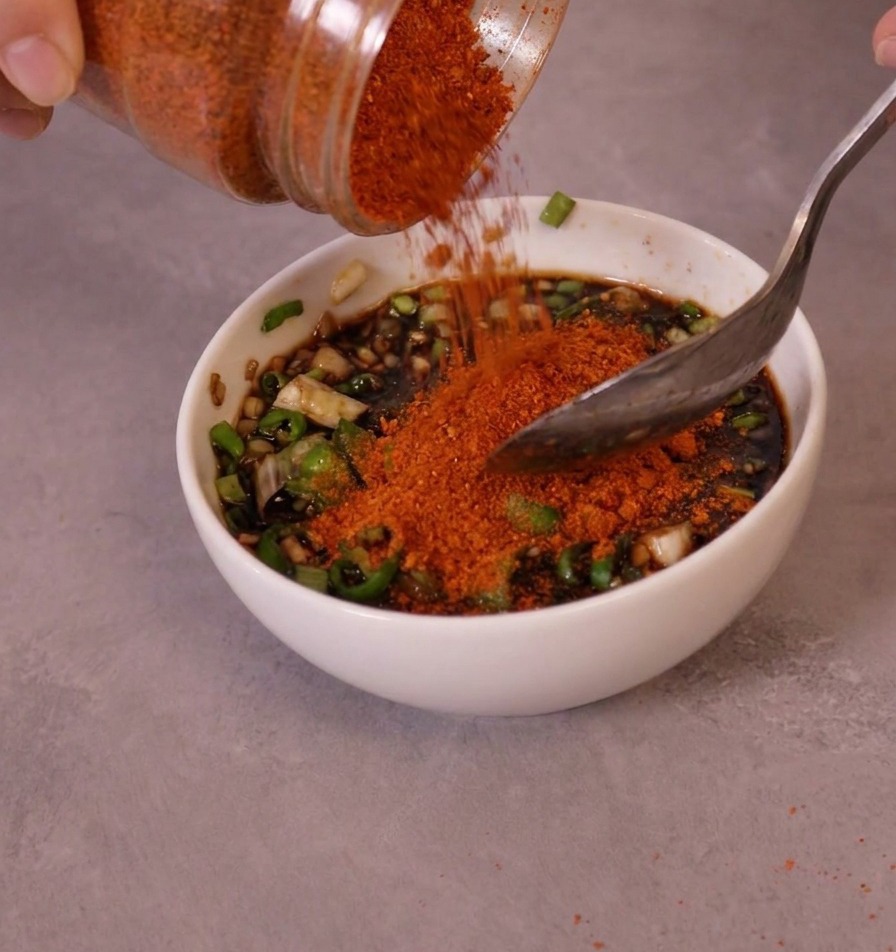 Leuchtend rotes, feines Chilipulver wird aus einem Glas in eine Schüssel mit der vorbereiteten Saucenbasis und grünen Chilis geschüttet.
