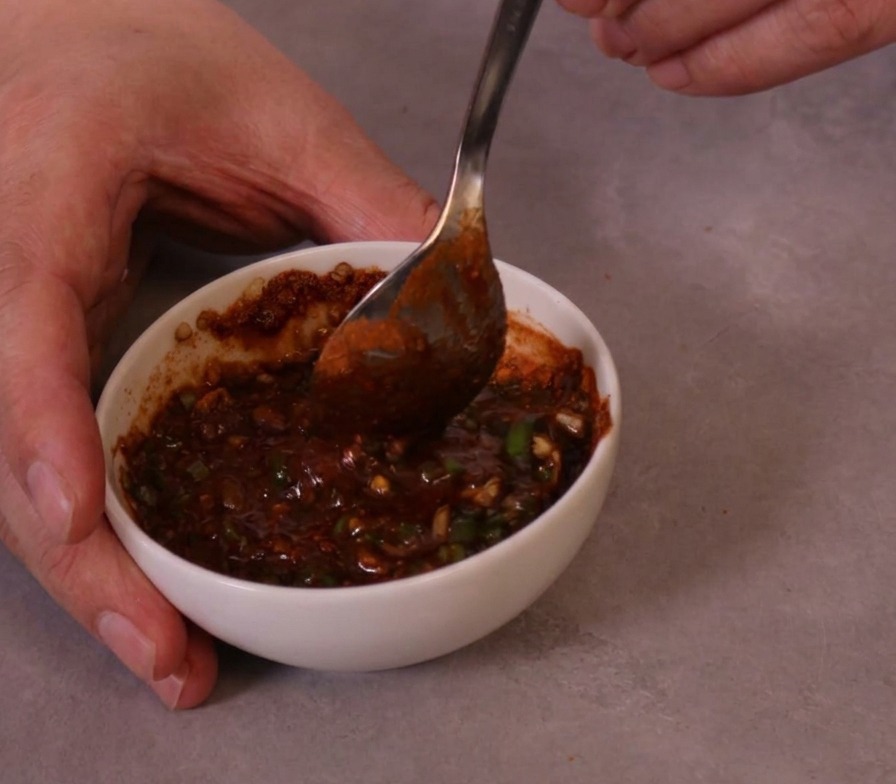 Eine dicke, leuchtend rote, scharfe Dip-Sauce wird mit einem Metalllöffel in einer kleinen Keramikschale umgerührt.