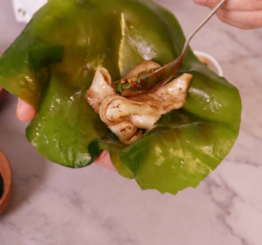 Eine Hand hält ein großes, leicht durchscheinendes grünes Kelp-Blatt, in dessen Mitte ein Stück fertig gegarter Schweinebauch liegt.