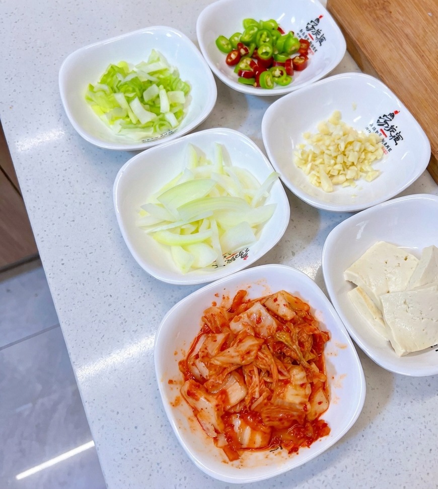 Kleine weiße Schälchen mit gehackten Frühlingszwiebeln, Peperoni, gehacktem Knoblauch, Zwiebelscheiben, Tofu und Kimchi.