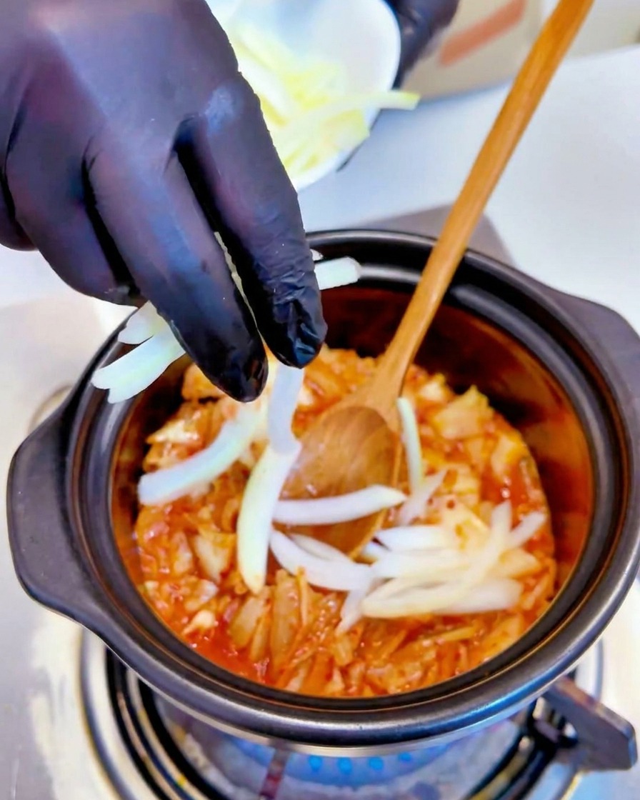 In Streifen geschnittene weiße Zwiebeln werden von einer Hand mit schwarzem Handschuh in den köchelnden Kimchi-Topf gegeben.