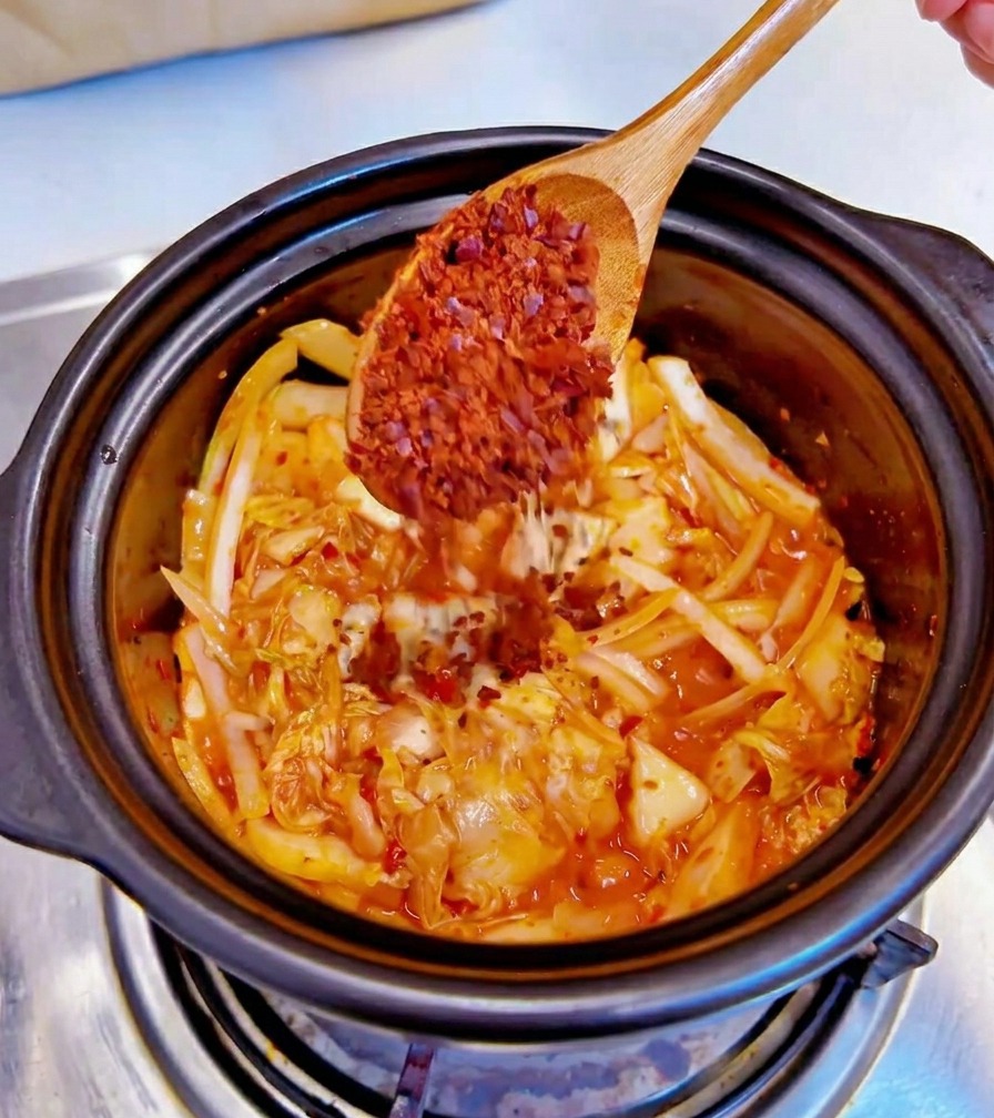 Ein Holzlöffel gibt grobe rote Chiliflocken in einen schwarzen Topf mit angebratenem Kimchi und Zwiebeln.