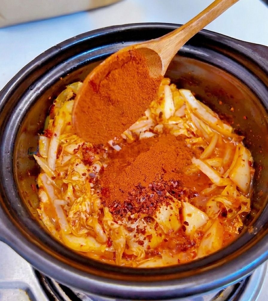 Feines rotes Chilipulver wird von einem Holzlöffel auf die gewürzte Kimchi-Zwiebel-Mischung gestreut.