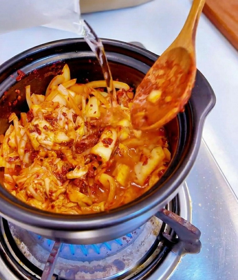 Wasser wird aus einem klaren Messbecher in einen schwarzen Topf mit scharfem Kimchi und Zwiebeln gegossen.