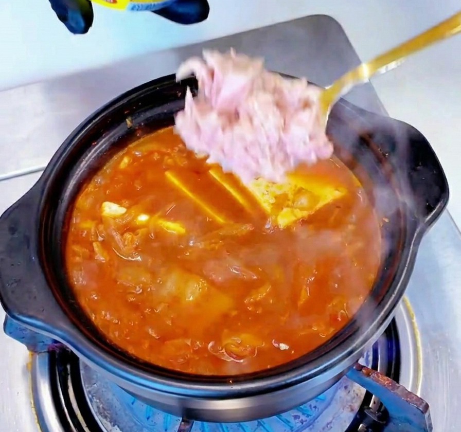 Stücke von hellrosa Thunfisch aus der Dose werden mit einem Löffel in die Mitte des köchelnden Eintopfs gegeben.