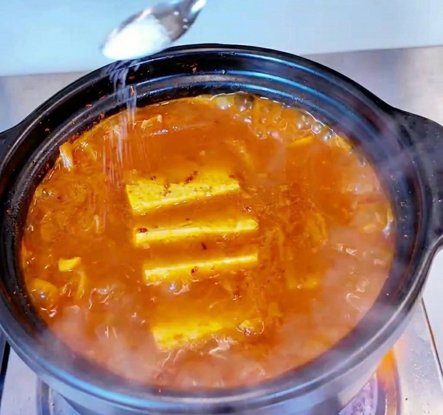 Ein kleiner Metalllöffel streut weißes Salz über die Oberfläche des brodelnden Kimchi-Eintopfs.