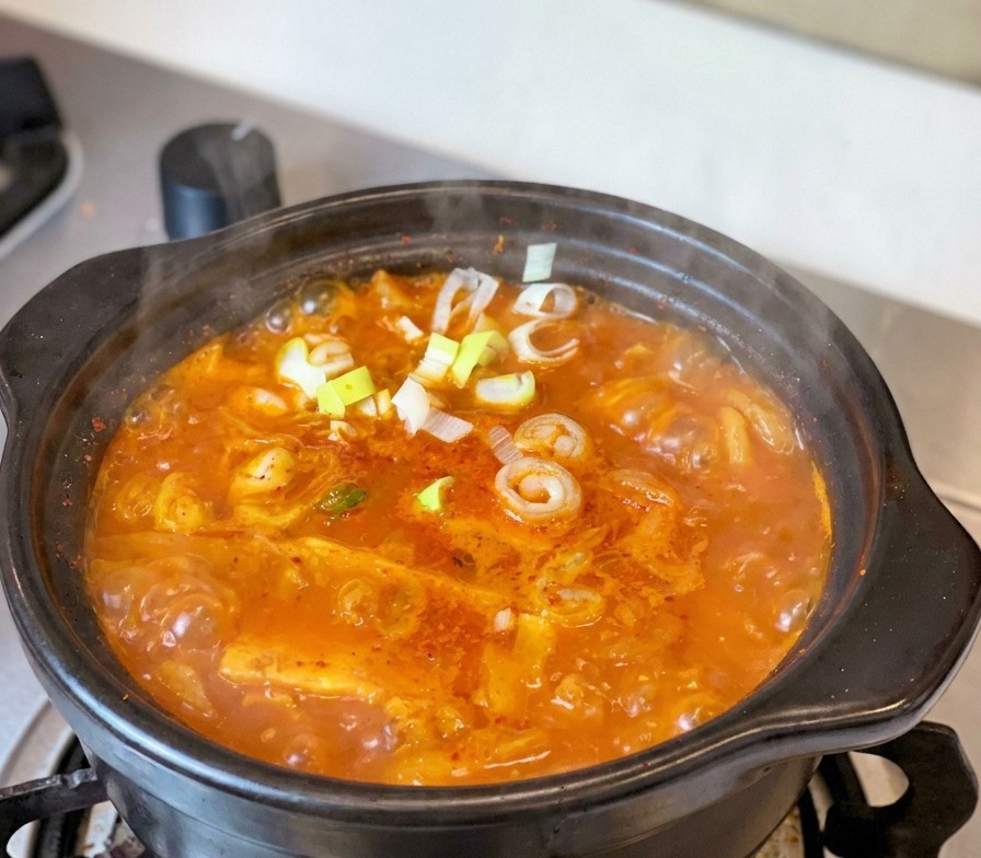 Ein brodelnder roter Kimchi-Eintopf mit Thunfisch in einem traditionellen schwarzen Tontopf, garniert mit frischen Frühlingszwiebeln und Peperonischeiben.