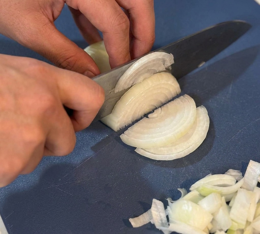 Hände schneiden eine weiße Zwiebel mit einem Kochmesser in feine Streifen auf einem blauen Schneidebrett.