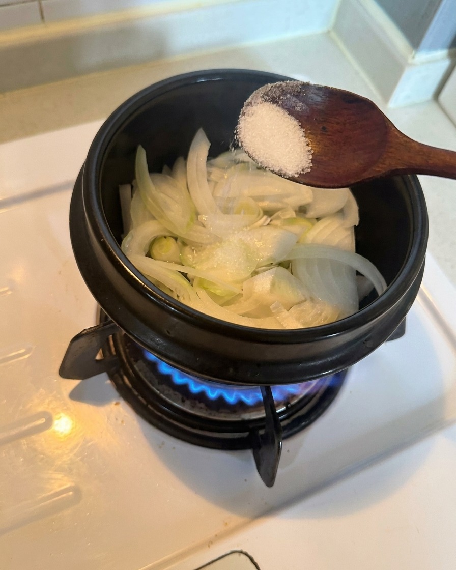 Geschnittene weiße Zwiebeln in einem Topf werden über einer brennenden Gasflamme mit einem Löffel weißem Zucker bestreut.