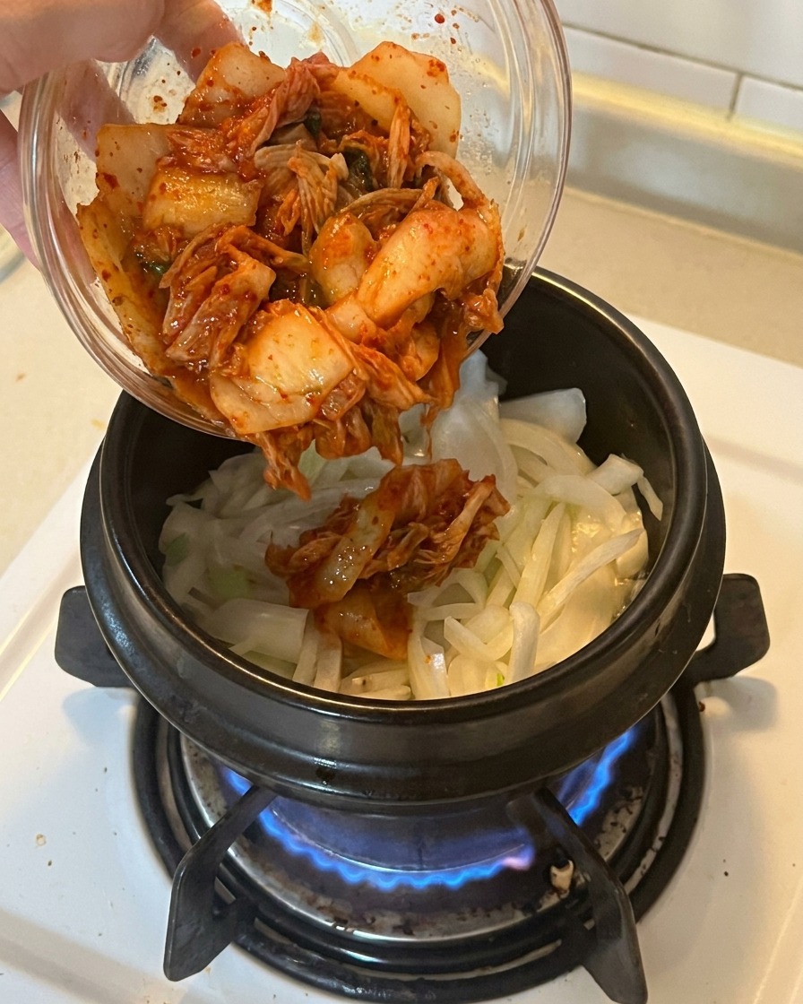 Eine Schale mit leuchtend rotem, gehacktem Kimchi wird in einen Topf mit angedünsteten Zwiebeln und Frühlingszwiebeln gegeben.