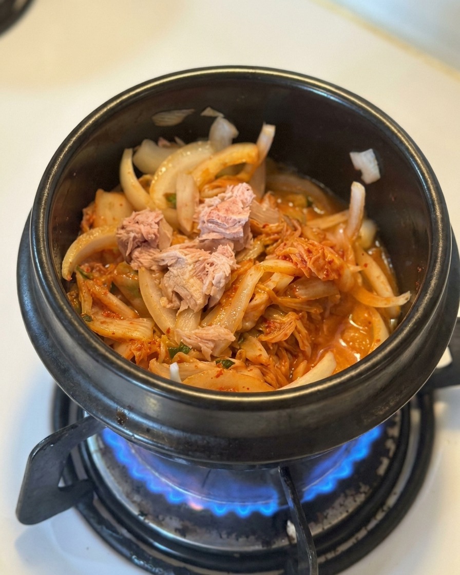 Stücke von Dosenthunfisch liegen auf einem Bett aus angeschwitztem Kimchi und Zwiebeln in einem schwarzen Topf.