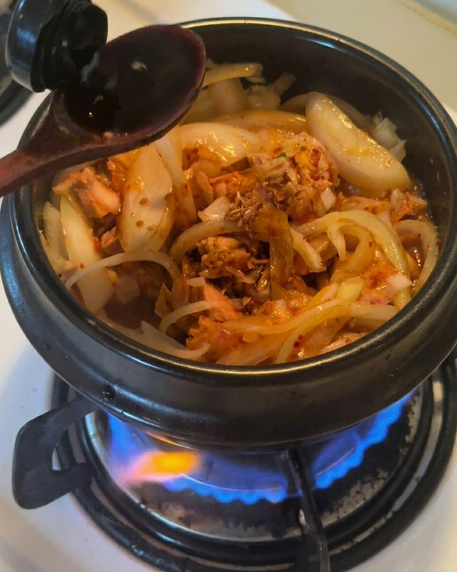 Ein Holzlöffel gießt dunkle Sojasauce in einen schwarzen Tontopf mit gebratenen Zwiebeln, Thunfisch und Kimchi.