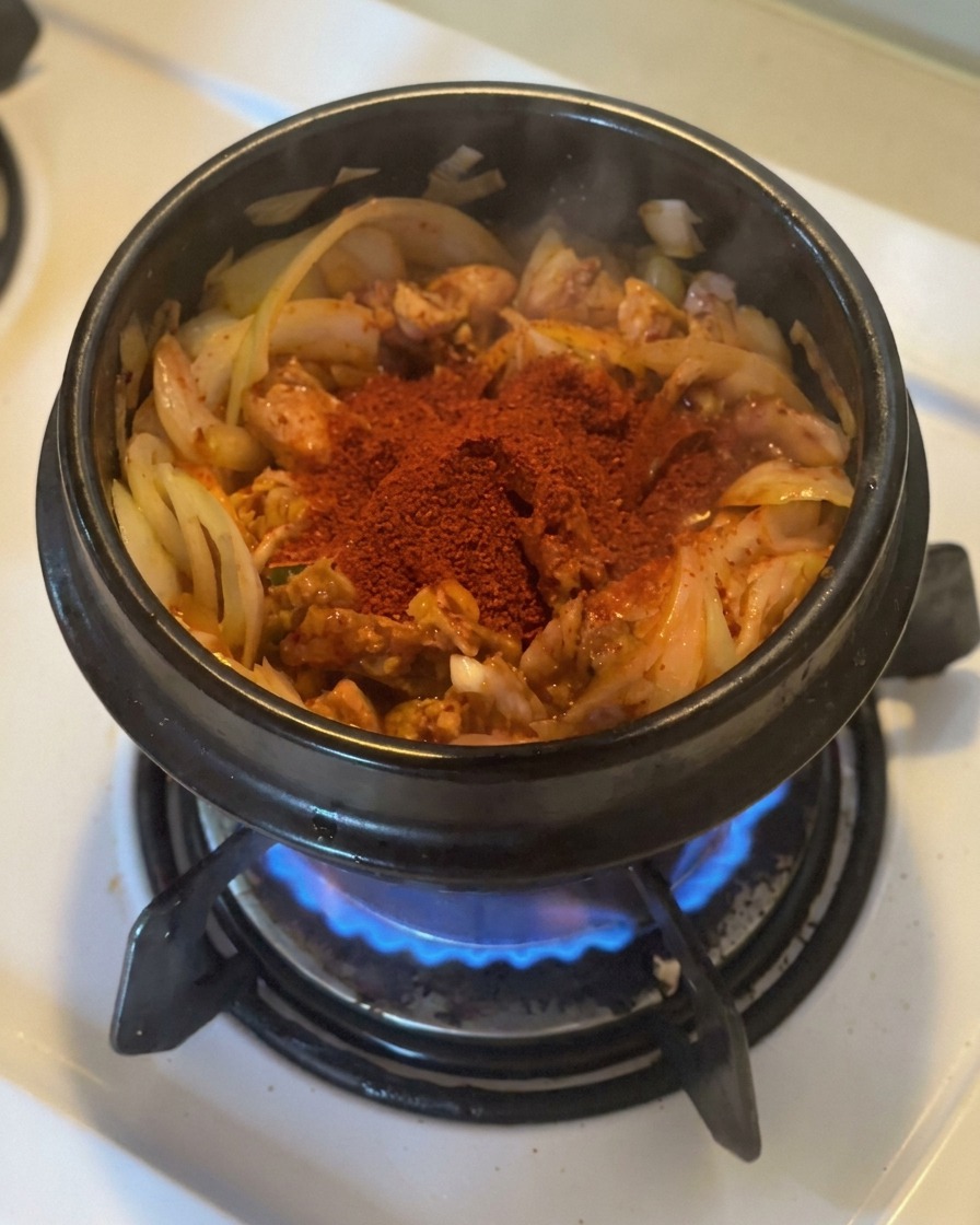 Ein großer Haufen leuchtend rotes Gochugaru liegt auf angeschwitztem Kimchi und Zwiebeln in einem schwarzen Steintopf über einer Gasflamme.