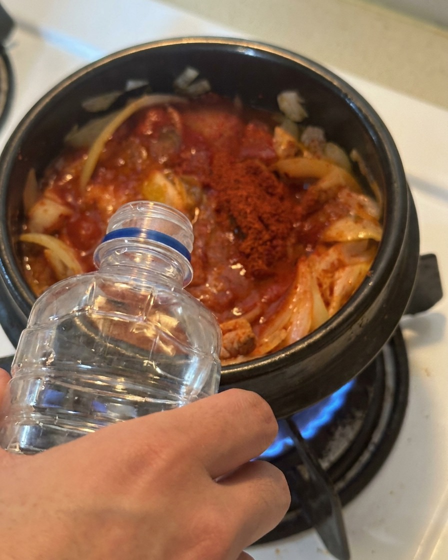 Wasser wird aus einer klaren Plastikflasche in einen Topf mit rot gewürztem Kimchi und Thunfisch gegossen.