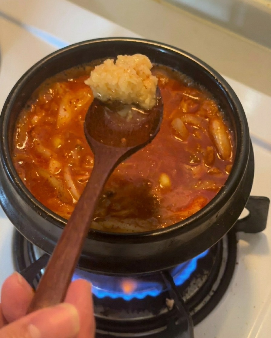 Ein Holzlöffel hält eine Portion feingehackten Knoblauch über einen köchelnden, roten Thunfisch-Kimchi-Eintopf.