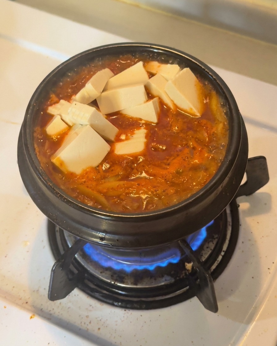 Frische weiße Tofuwürfel und geschnittene grüne Chilischoten sind dekorativ auf einem kochenden, roten Kimchi-Eintopf in einem schwarzen Tontopf angerichtet.