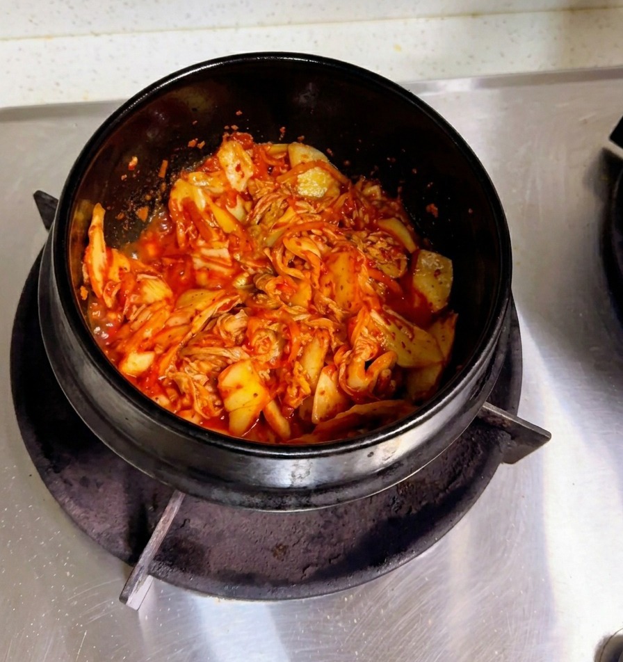 Eine Hand gießt trübes Reiswasser in einen Steintopf mit angebratenem Kimchi.