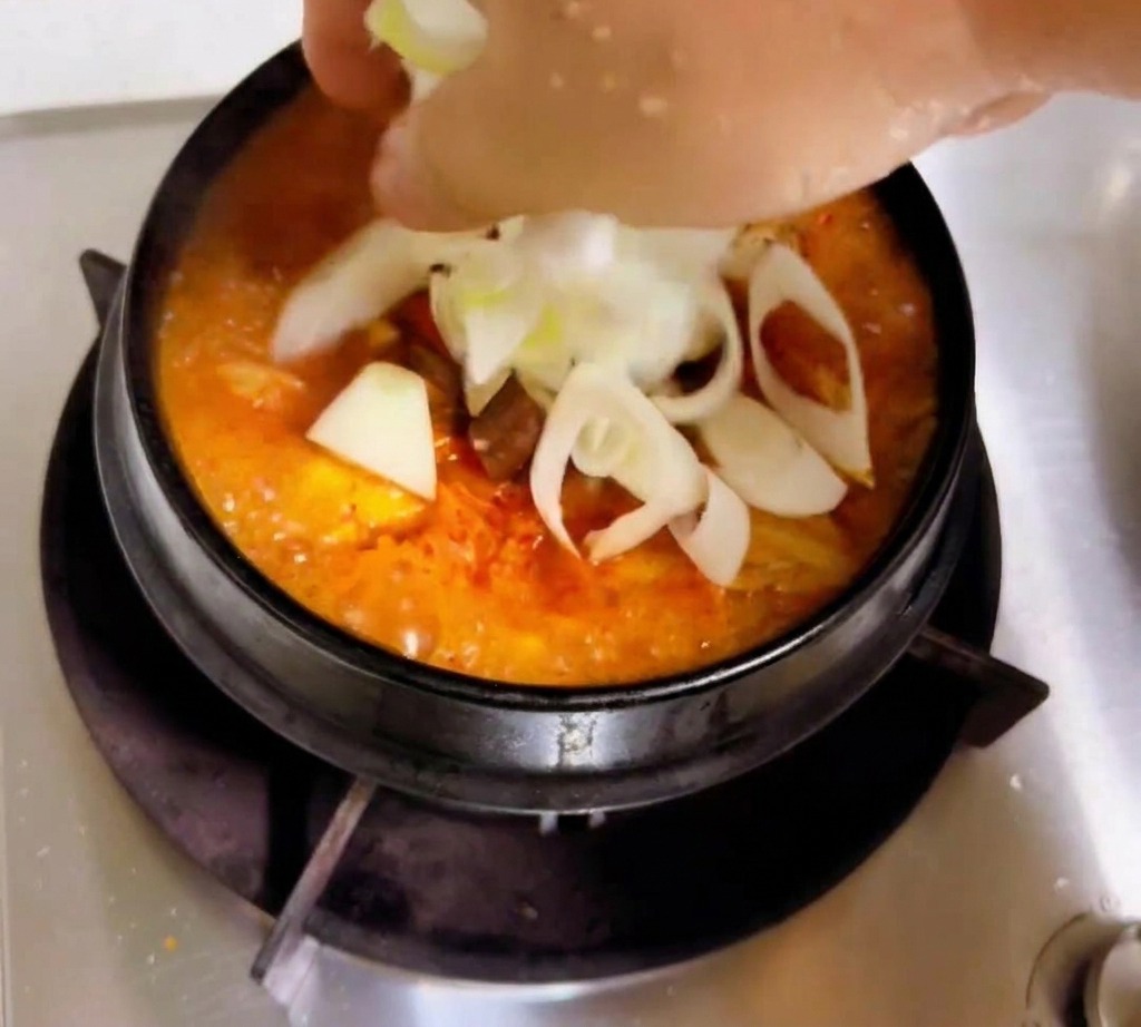 Eine Hand gibt geschnittene weiße Zwiebeln und Frühlingszwiebeln in den köchelnden orange-roten Eintopf.