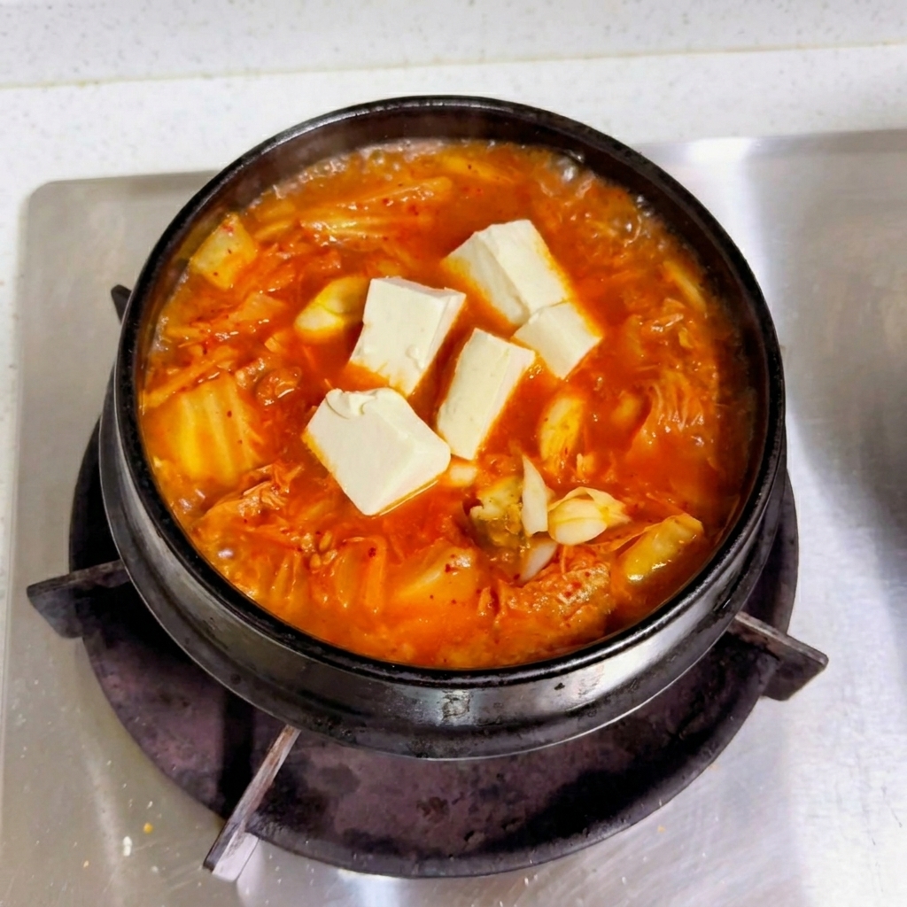 Frische weiße Tofuwürfel liegen auf einer kochenden roten Kimchi-Brühe in einem schwarzen Topf.