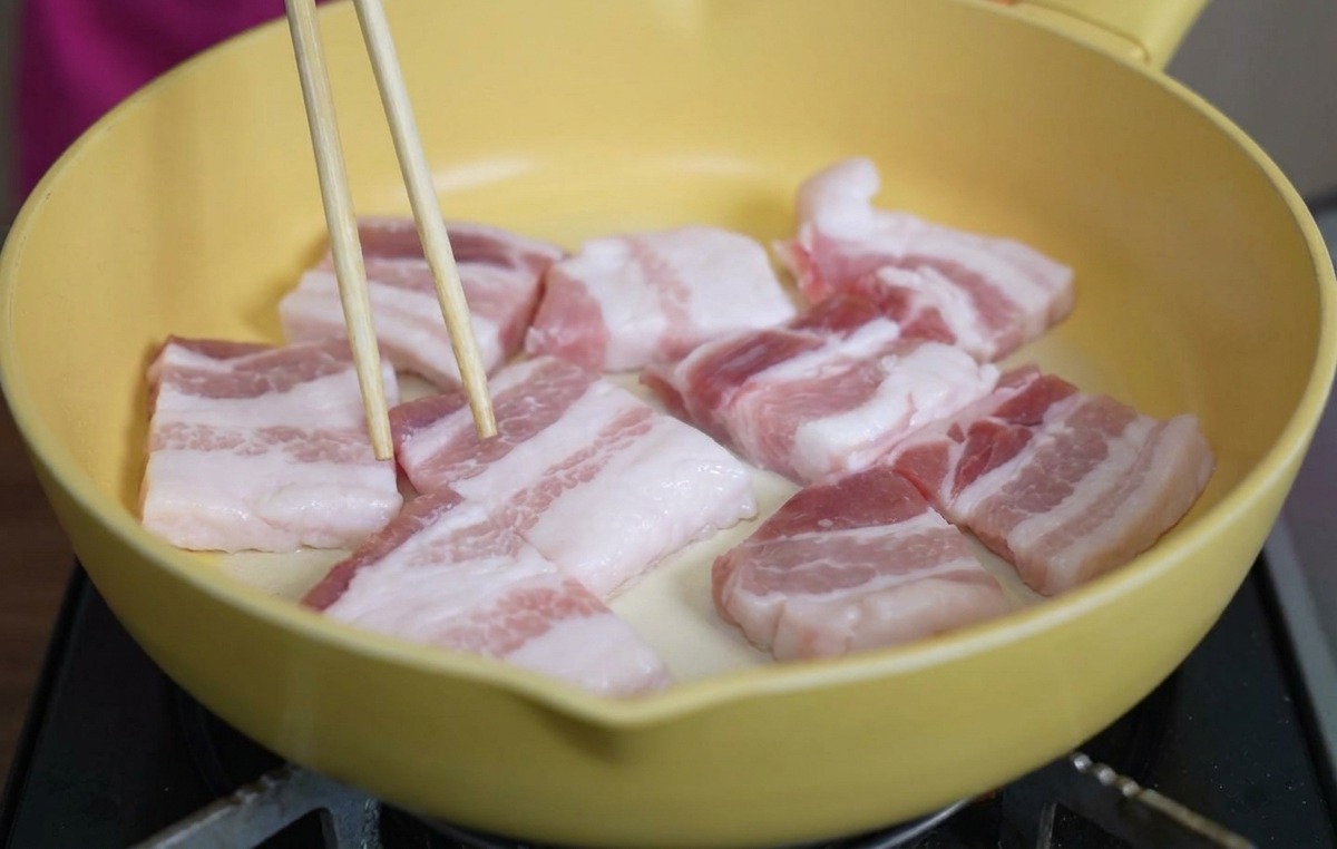 Rohe, dicke Scheiben von Schweinebauch in einer gelben Keramikpfanne, die mit Stäbchen gewendet werden.