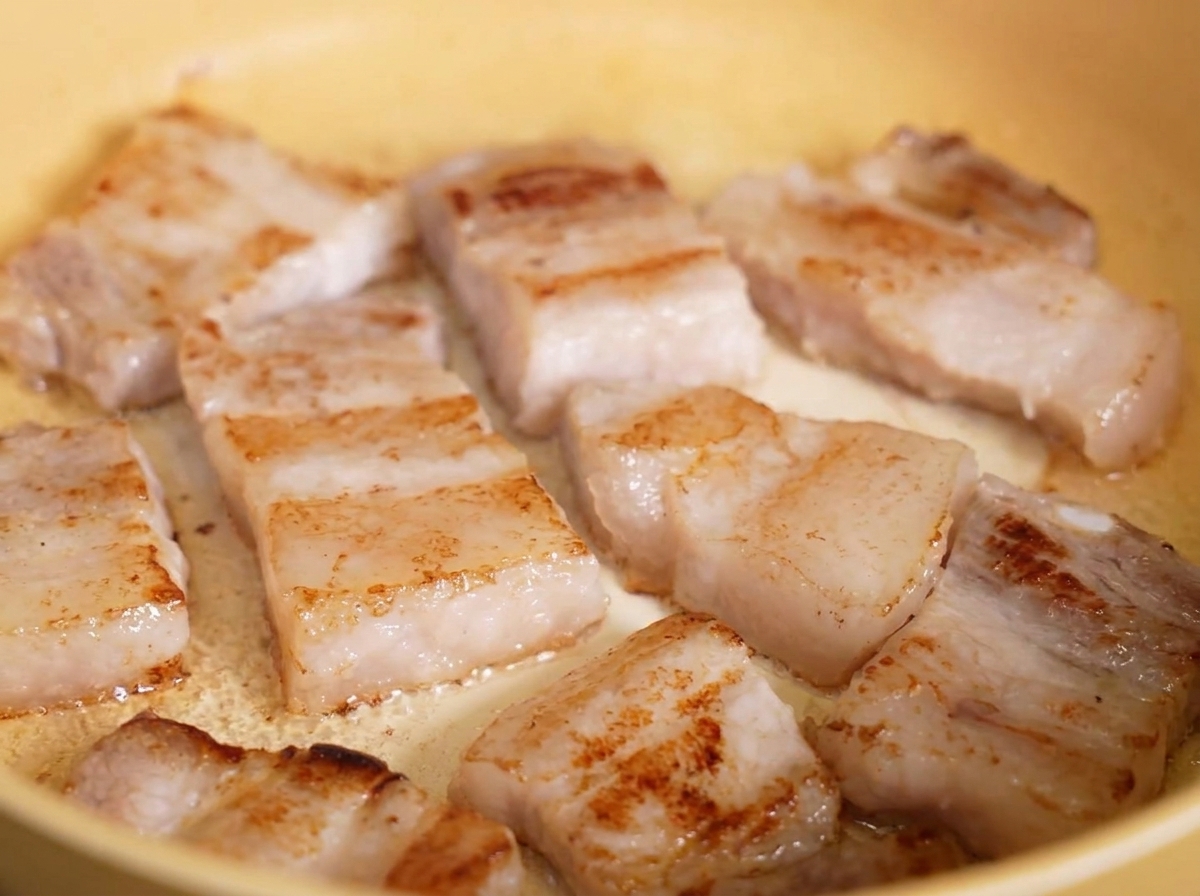 Knusprig goldbraun gebratene Schweinebauchscheiben, die im eigenen Fett in einer gelben Pfanne brodeln.