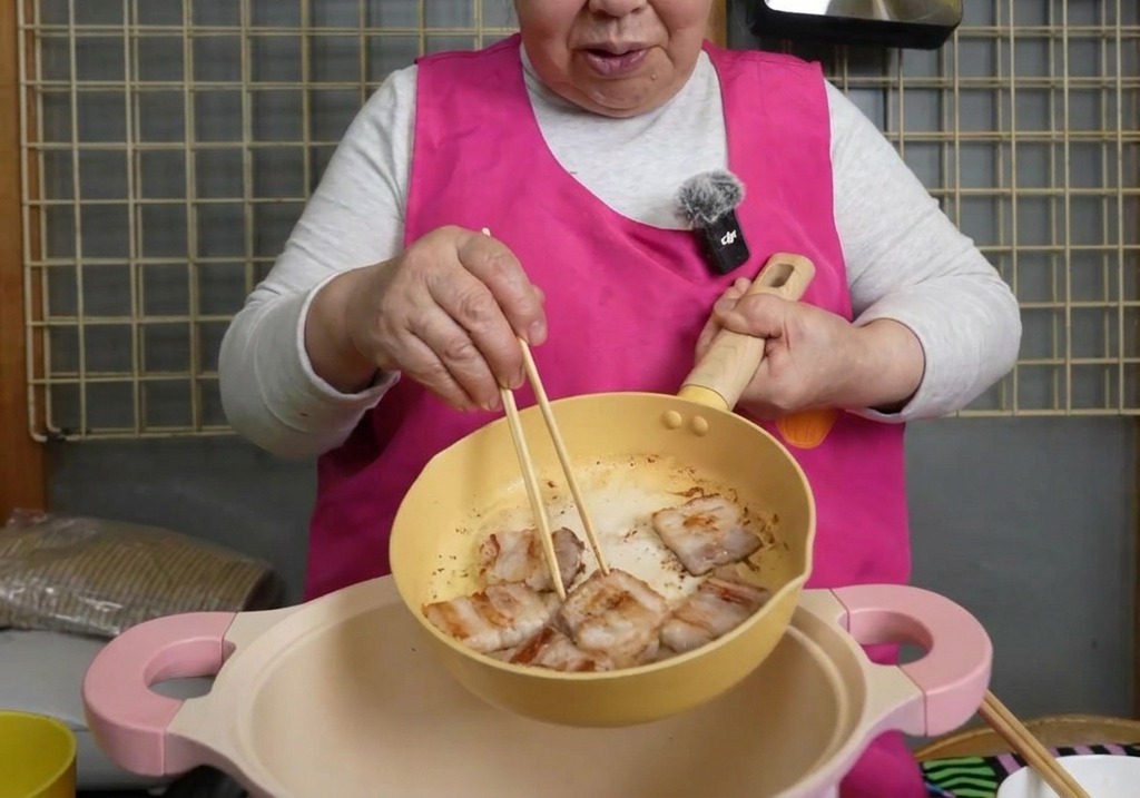Gebräunte Schweinebauchscheiben werden mit Stäbchen aus einer gelben Pfanne in einen größeren rosa Topf gelegt.
