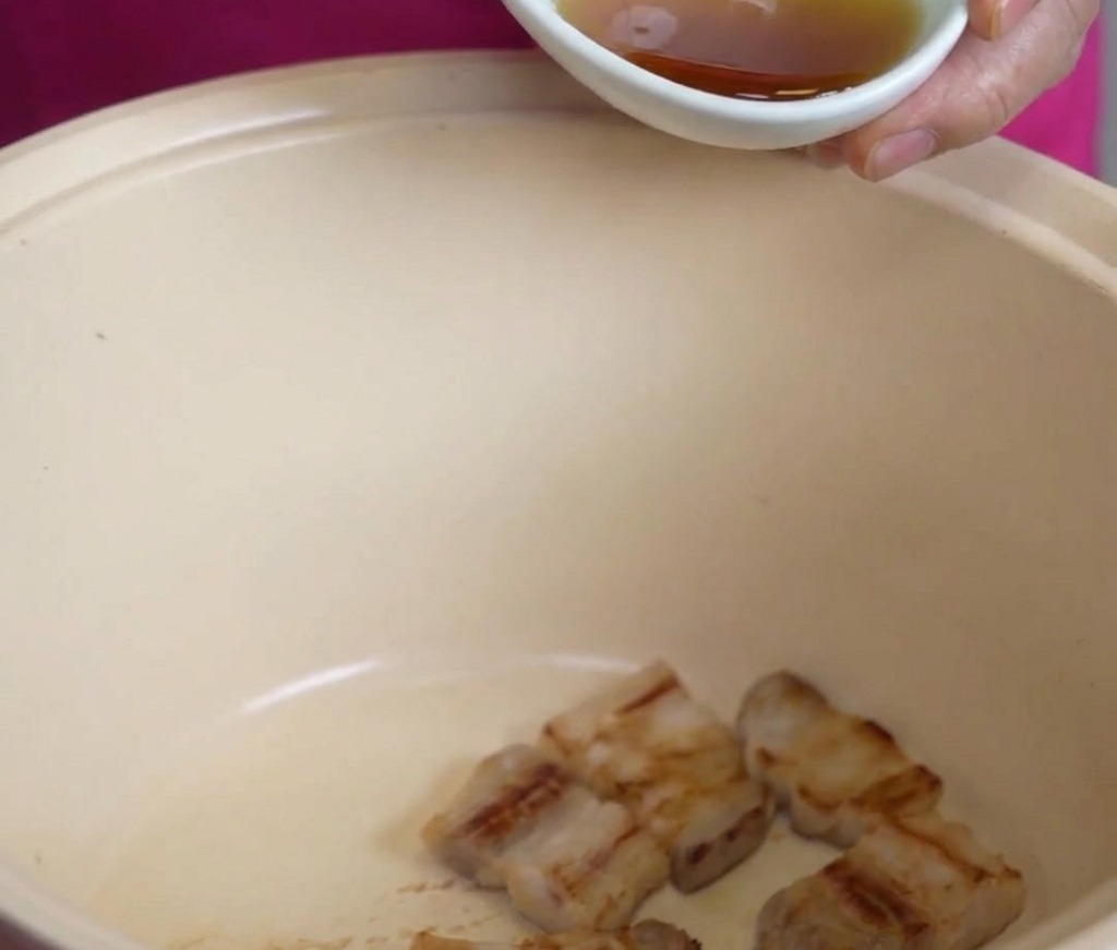 Aus einer kleinen weißen Schüssel wird goldenes Sesamöl über gebratene Schweinebauchstücke in einem großen cremefarbenen Topf gegossen.