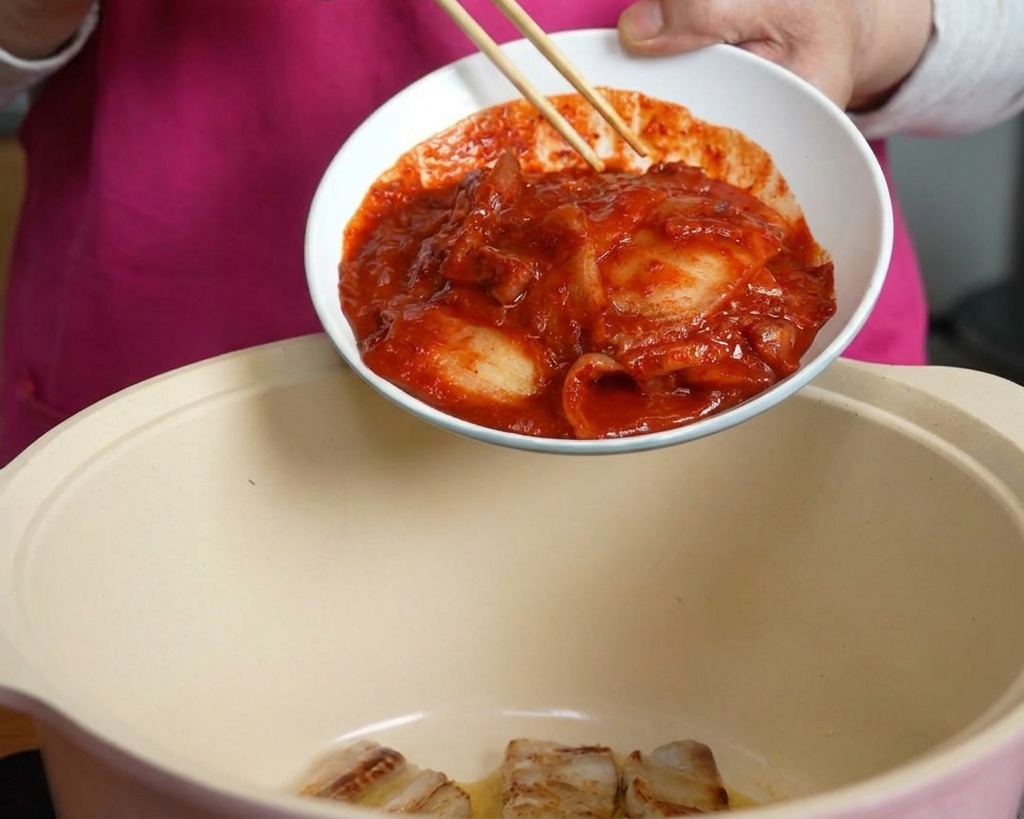 Eine Hand schiebt mit Stäbchen eine Schüssel mit scharfem rotem Kimchi in einen Kochtopf mit Schweinebauch.