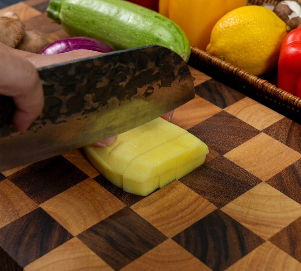 Ein Messer schneidet eine geschälte gelbe Kartoffel auf einem Holzschneidebrett in gleichmäßige Würfel.