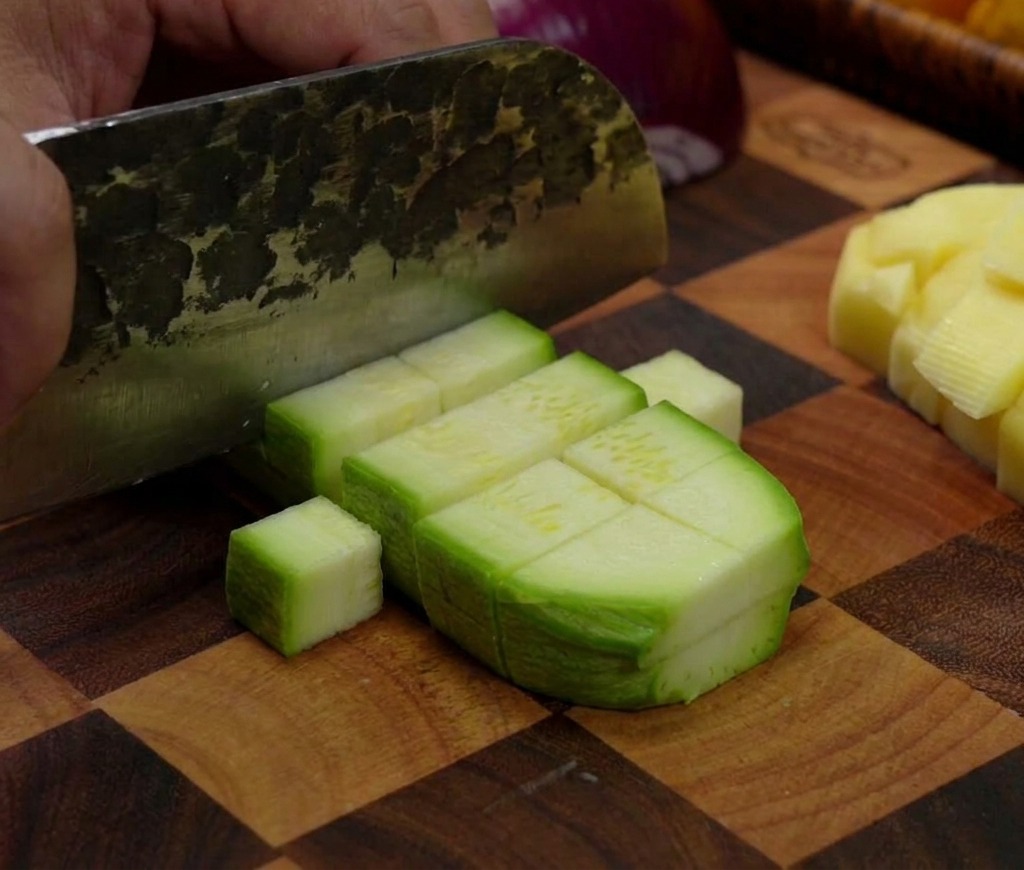 Ein Hackmesser schneidet eine grüne Zucchini auf einem Holzbrett in kleine, rechteckige Stücke.